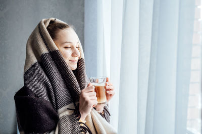 Cozy mood, baby its cold outside. young woman standing near window with plaid, drinking coffee or