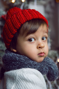 Child in a white sweater and a red knitted hat sits on the steps with snow at home at christmas