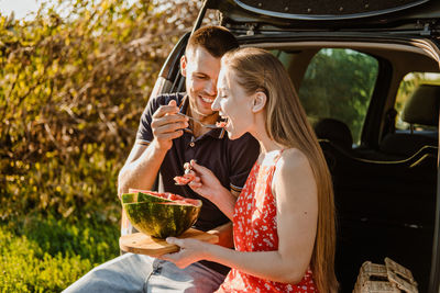 Millennial couple sitting on open trunk and eating watermelon. happy young couple having break at