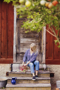 Full length of woman cutting radishes while sitting on steps outside wooden cottage