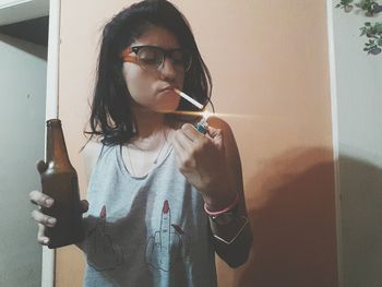 Close-up of young woman holding drink while igniting cigarette at home