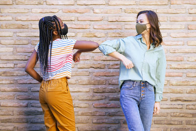 Smiling friends wearing mask doing elbow bump against wall