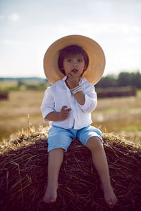 Baby boy in straw hat and blue pants sitting on a haystack in a field in autumn