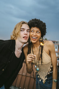 Young man sticking out tongue by female friend holding lit sparkler on rooftop
