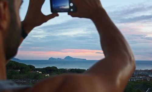 Midsection of man photographing sea against sky during sunset