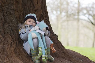 Portrait of cute girl holding toy while sitting by tree trunk during winter