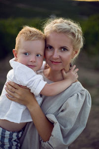 Blonde woman in a dress holds her baby boy in her arms in a vineyard during sunset