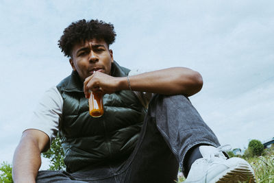 Low angle portrait of young man drinking juice from bottle against sky