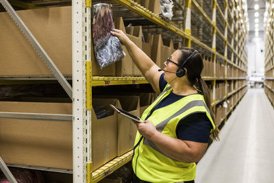 Mature female worker with digital tablet checking merchandise on rack while talking through headset at distribution ware