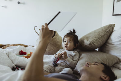 Father reading book with baby son lying on bed at home