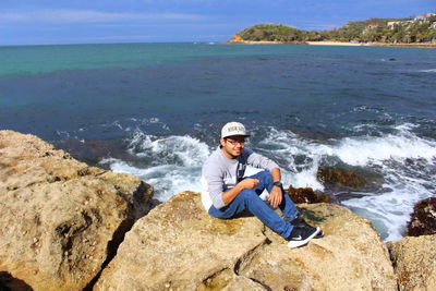 Man sitting on rock at sea shore against sky