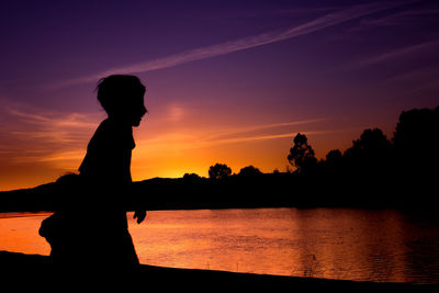 Silhouette boy standing by lake against sky during sunset