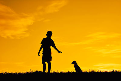 Silhouette boy with dog on field against sky during sunset