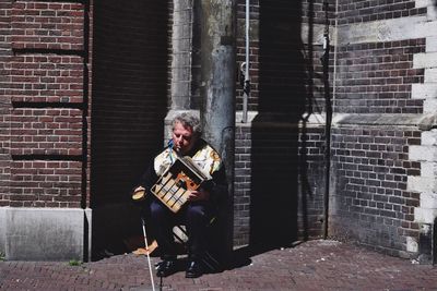 Full length of man holding box while sitting on chair by brick wall