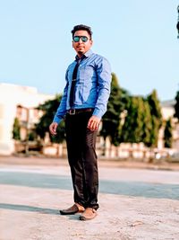 Portrait of young man wearing sunglasses standing on retaining wall against clear sky