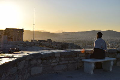 Rear view of man sitting at observation point against clear sky during sunset