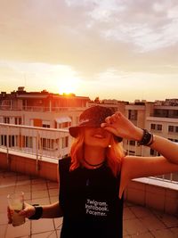 Woman holding drink while standing on terrace in city against sky