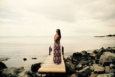 Full length of woman standing on jetty leading towards sea against cloudy sky