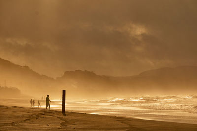 Silhouette people walking at beach against orange sky during sunrise