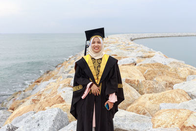 Portrait of young woman in graduation gown standing at beach against cloudy sky