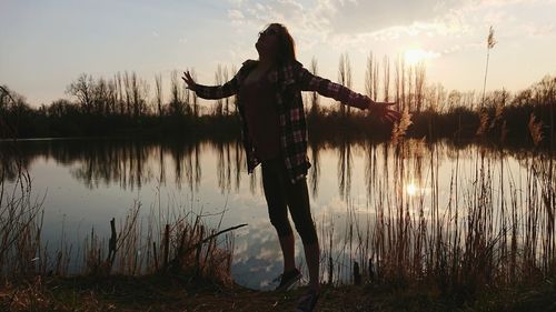 Woman with arms outstretched standing at lake against sky during sunset