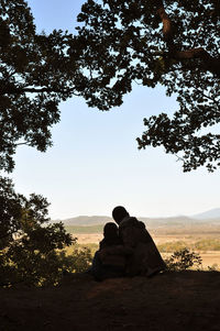 Rear view of silhouette man sitting on rock against sky