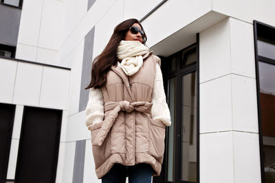 Stylish model with long brunette hair wearing knitted white scarf and sweater, beige down vest