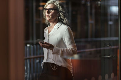 Thoughtful senior businesswoman wearing headphones while using mobile phone at office