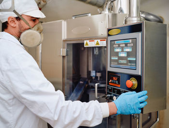 Side view of male chemist in uniform operating capsule filling machine at pharmaceutical manufacturing plant