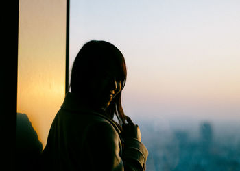 Woman standing by window during sunset