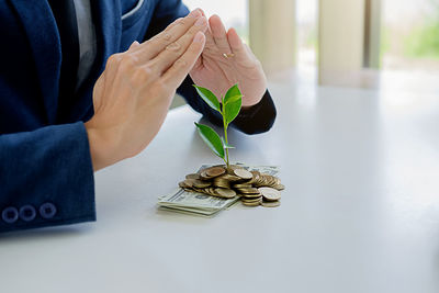 Midsection of man shielding coins with plant and paper currency on table