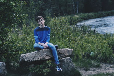 Full length of thoughtful teenage boy sitting on rocks against lake