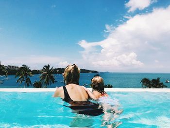 Rear view of woman with daughter swimming in pool by sea against cloudy sky