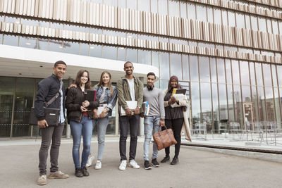 Full length portrait of smiling multi-ethnic students standing against building in campus at university