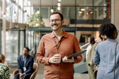 Portrait of smiling male business professional holding coffee cup and folder while standing amidst delegates at conventi