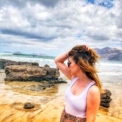 Young woman with hand in hair standing at beach against cloudy sky