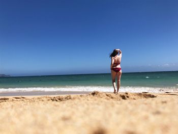 Rear view of woman in bikini walking towards shore at beach against blue sky