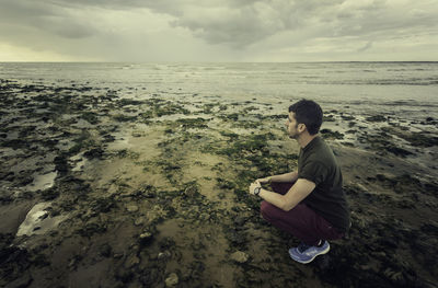 Side view of young man crouching at beach against cloudy sky