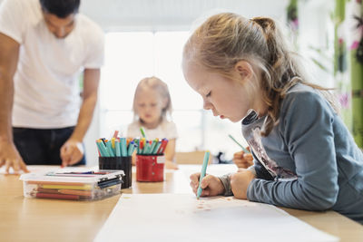 Girl using felt tip pen in drawing class with classmate and teacher in background