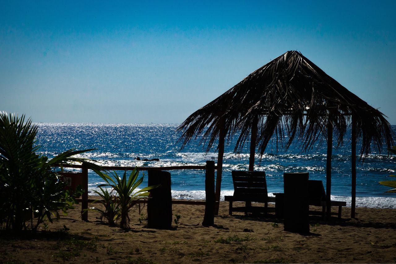 sky, ocean, beach, sea, land, water, nature, thatched roof, beauty in nature, chair, sand, scenics - nature, coast, horizon, tranquility, blue, travel destinations, tranquil scene, hut, horizon over water, body of water, clear sky, no people, tropical climate, seat, evening, vacation, holiday, trip, tree, architecture, shore, outdoors, bay, idyllic, plant, dusk, day, relaxation, travel, sunlight, parasol, summer, roof, sunset, palm tree, beach umbrella, beach hut, shade, tourism, sunny