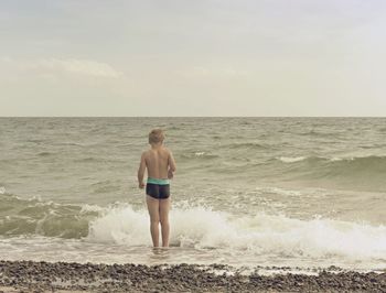 Blond hair boy stay in cold sea tide. kid on stony beach with foamy waves. windy day at  seascape