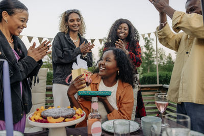 Cheerful friends applauding smiling woman holding gifts during birthday celebration in back yard