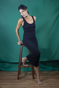 Portrait of adult model sitting on wooden chair against dark green background. 