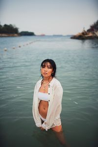 Portrait of young woman wearing bikini standing in sea against clear sky