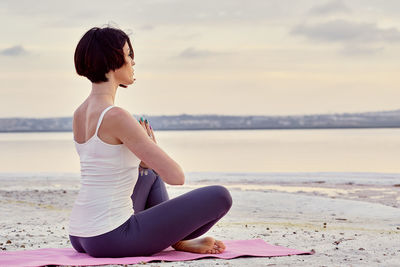 Side view of woman meditating while sitting at beach during sunset