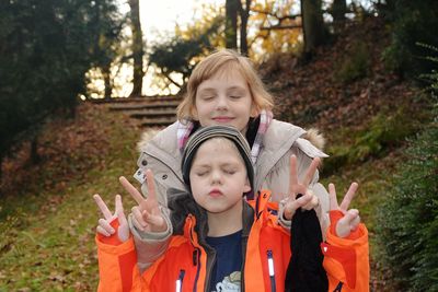 Siblings with eyes closed gesturing peace sign at park during sunset