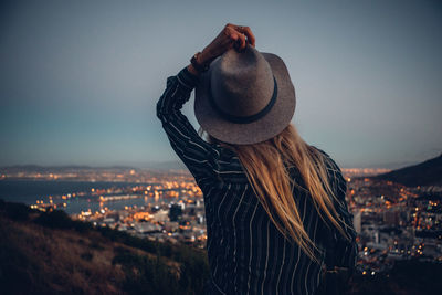 Rear view of woman by illuminated cityscape against clear sky during dusk