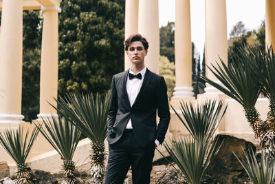 A beautiful young man, the groom in an elegant wedding suit, stands posing in the city's old park