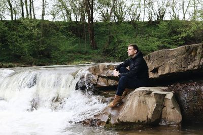 Thoughtful man sitting on rock by waterfall in forest