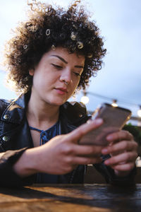 Young curly haired woman text messaging through smart phone sitting at table